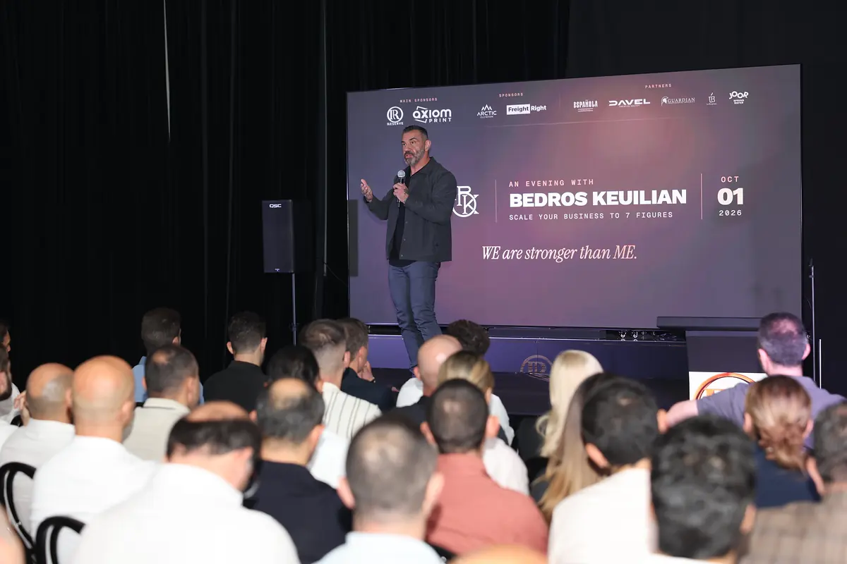 Bedros Keuilian speaking in front of the event backdrop screen, the audience in the foreground